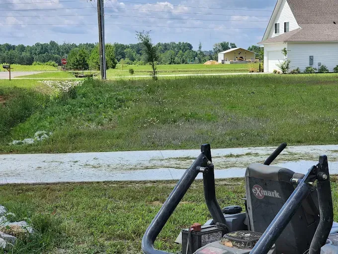 A riding lawnmower partially visible, cutting grass near a house in a rural setting.