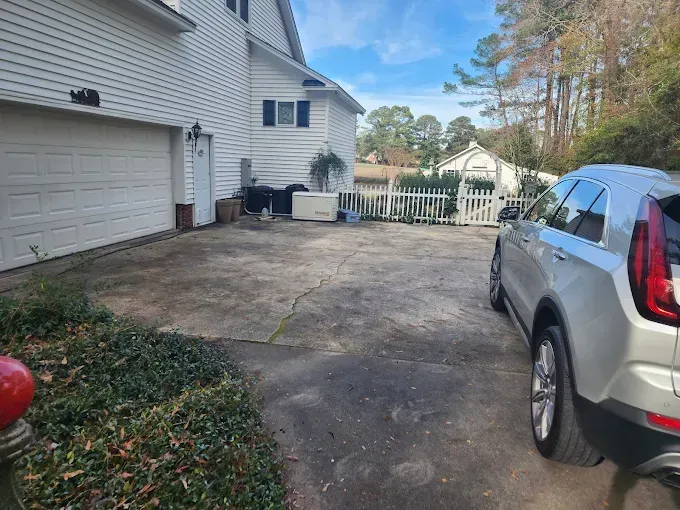 A silver SUV parked on a cracked driveway next to a white house with a garage and fence.