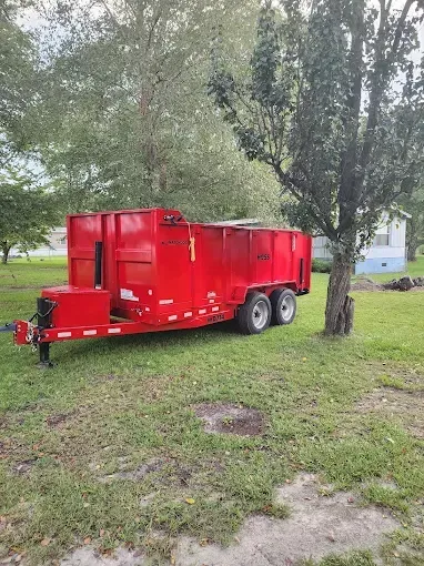 Red dump trailer parked on grass next to a tree.