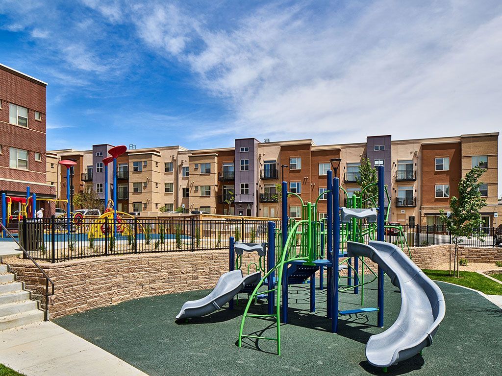 Playground equipment with slides on a rubber surface in front of multi-story brick and tan apartment buildings.