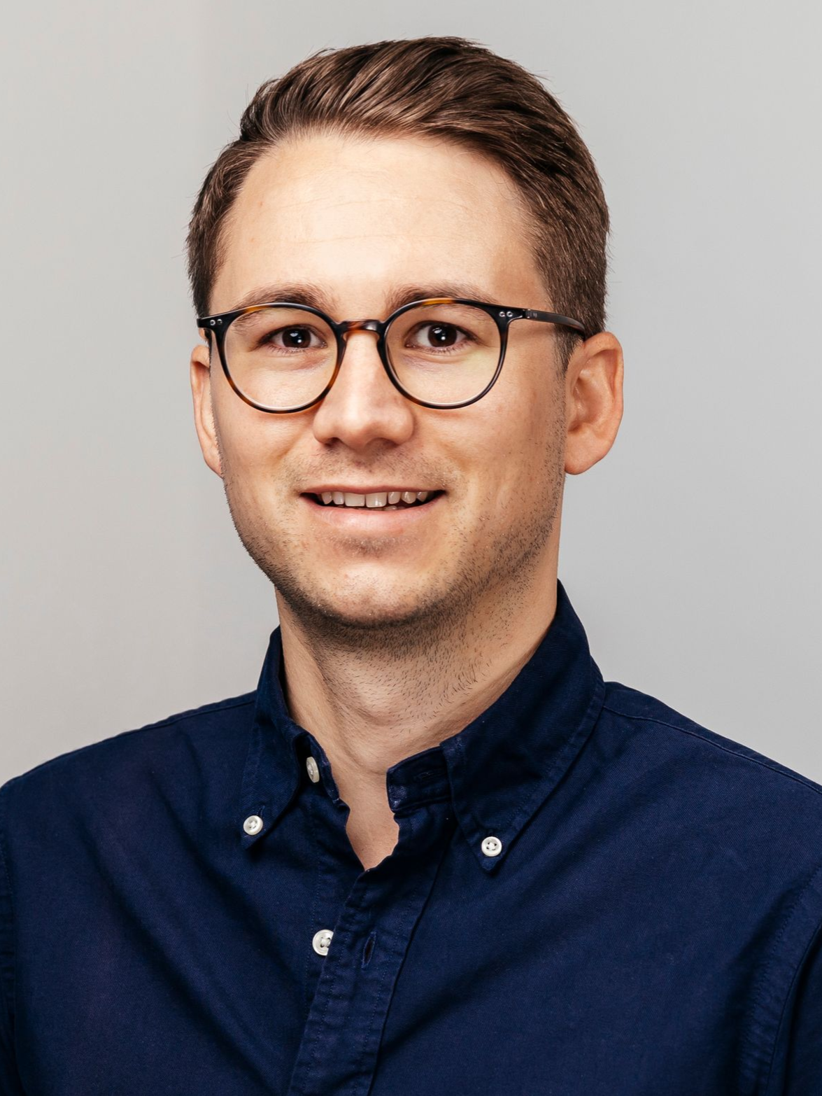 Mann mit Brille lächelt und trägt ein dunkelblaues Button-Down-Hemd vor grauem Hintergrund.