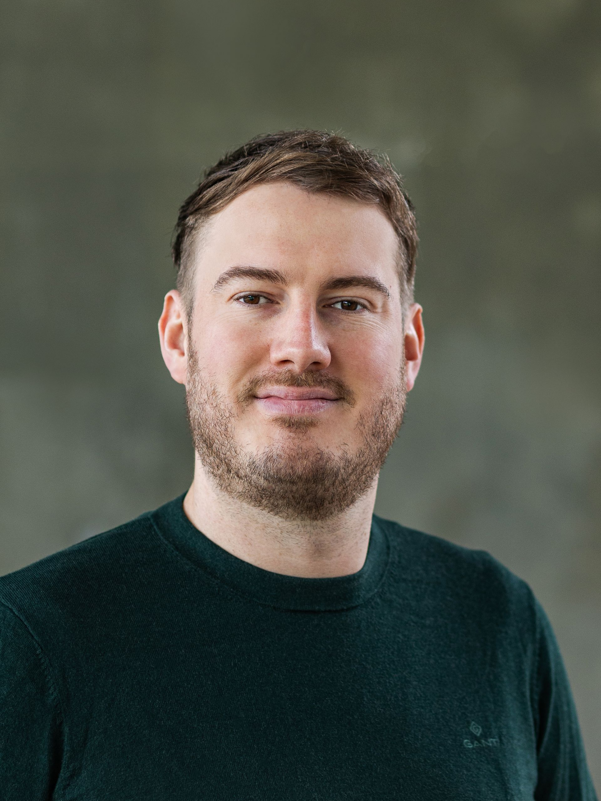 Mann mit braunem Haar und Bart, trägt einen grünen Pullover und lächelt vor einem grauen Hintergrund.