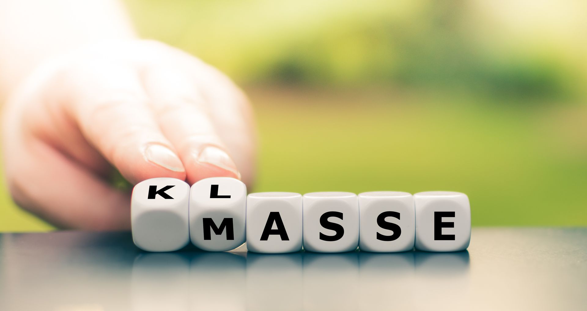 Von Hand angeordnete Buchstabenwürfel mit der Aufschrift „KL/MASSE“ vor einem verschwommenen grünen Hintergrund.