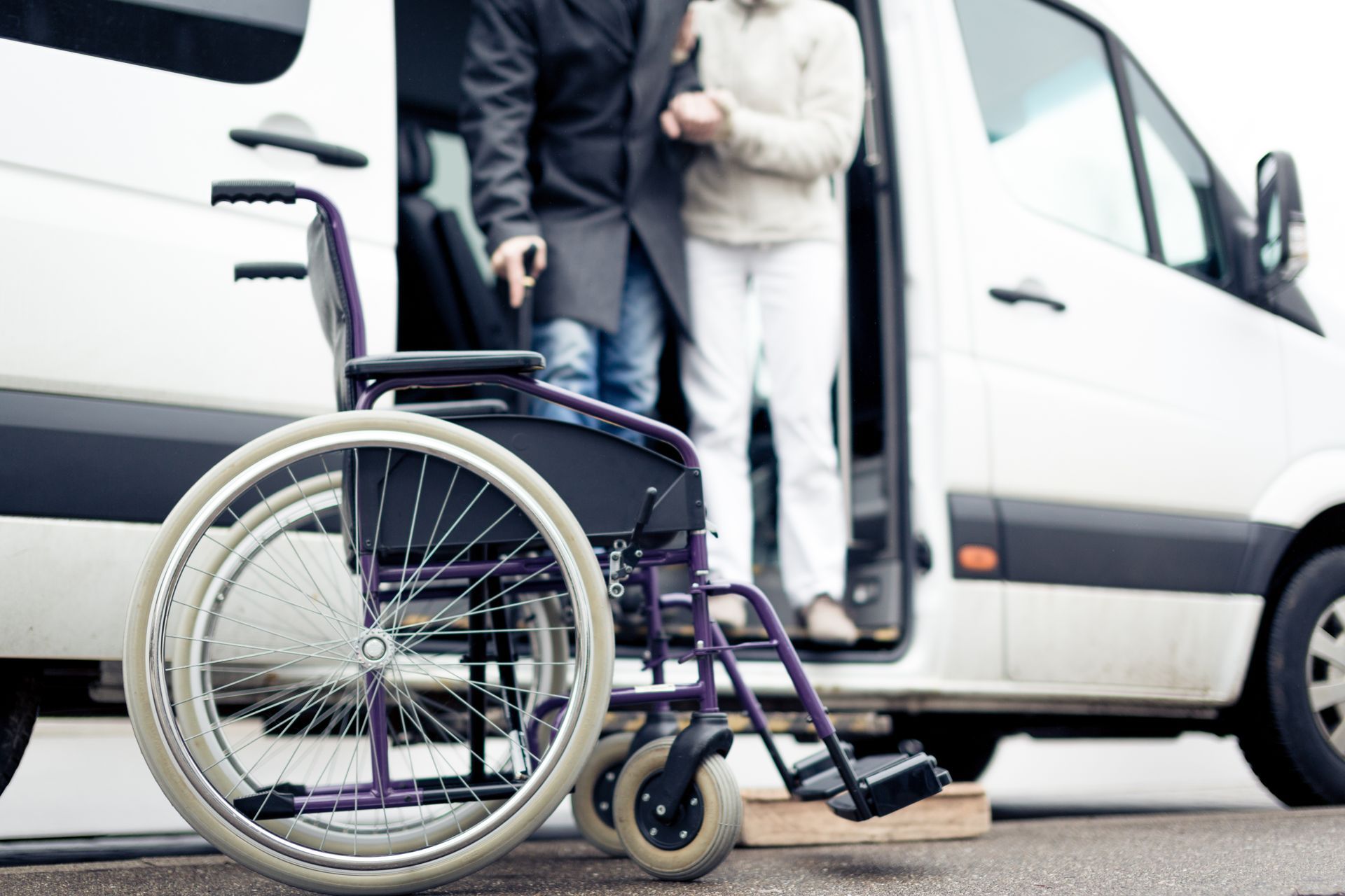 Rollstuhl vor einem weißen, barrierefreien Lieferwagen; Person wird beim Aussteigen aus dem Fahrzeug unterstützt.