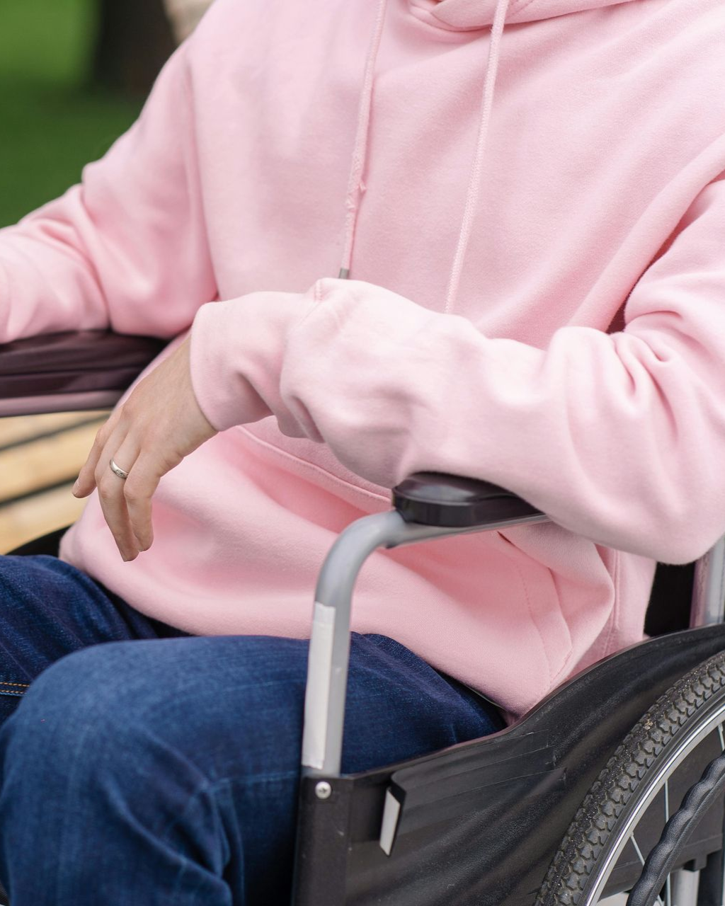 Person in einem rosa Kapuzenpullover, die im Freien in einem Rollstuhl sitzt und Blue Jeans trägt.