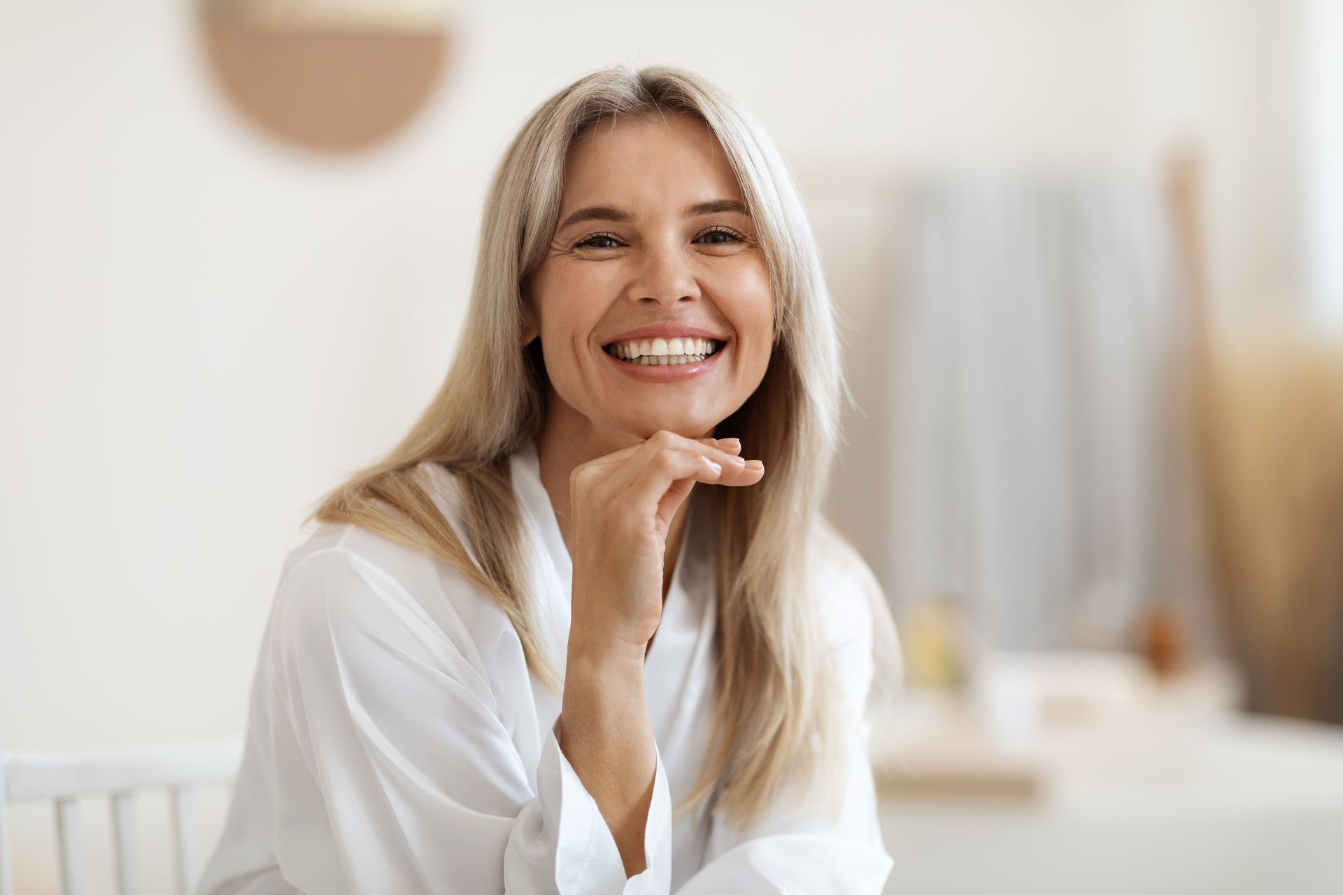 A smiling woman is sitting at a table with her hand on her chin.