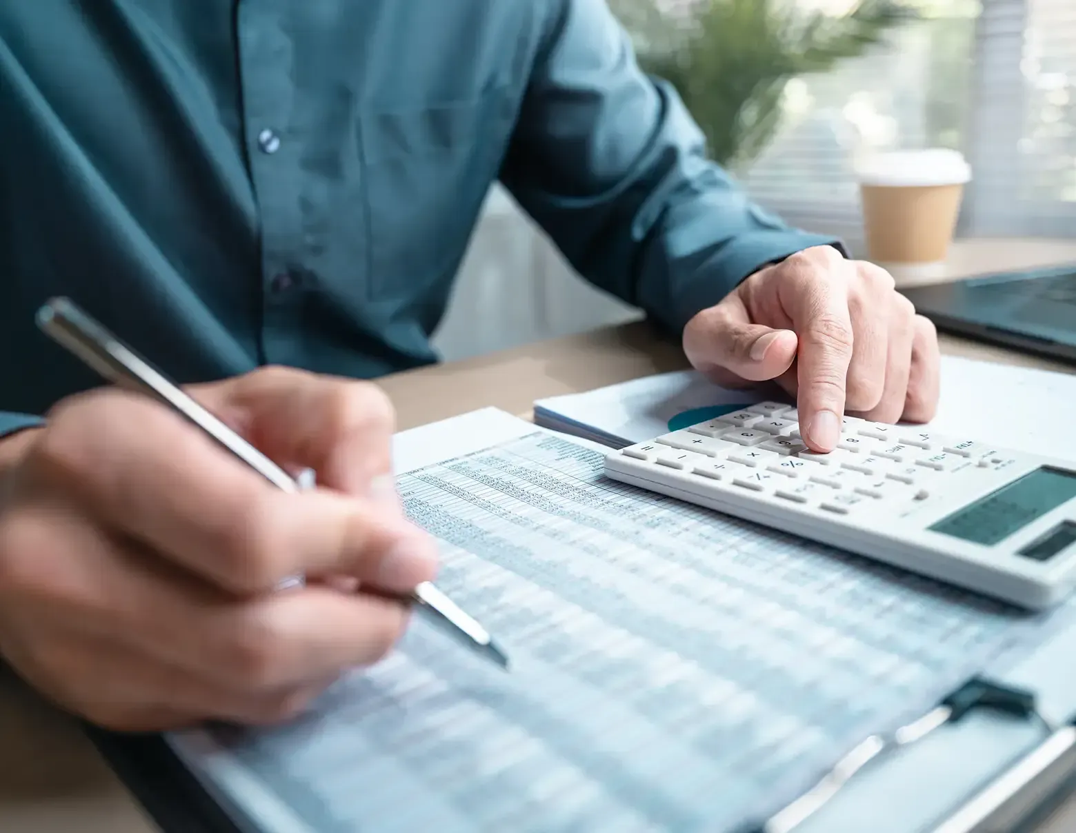 A person in a dark shirt uses a calculator and writes on a document at a desk with a coffee cup in the background.