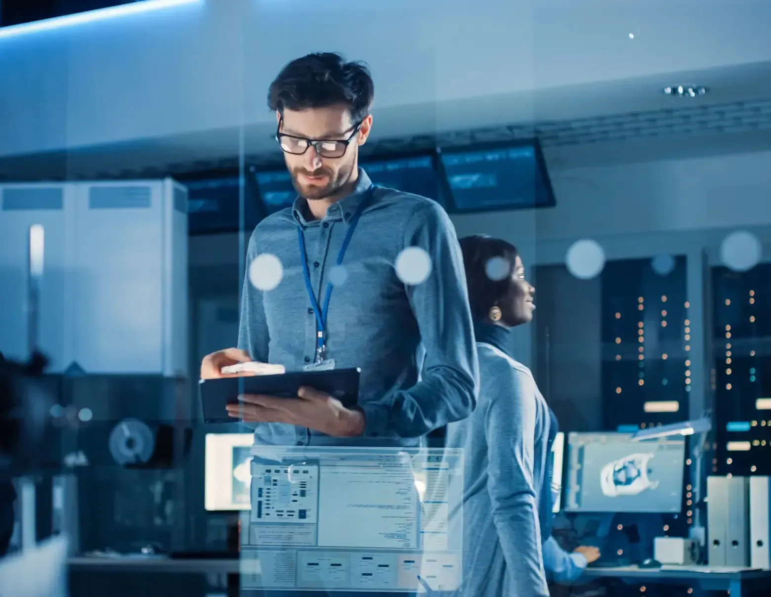 Man with glasses looks at tablet, in a tech lab with a second person at a computer. Blue lighting.