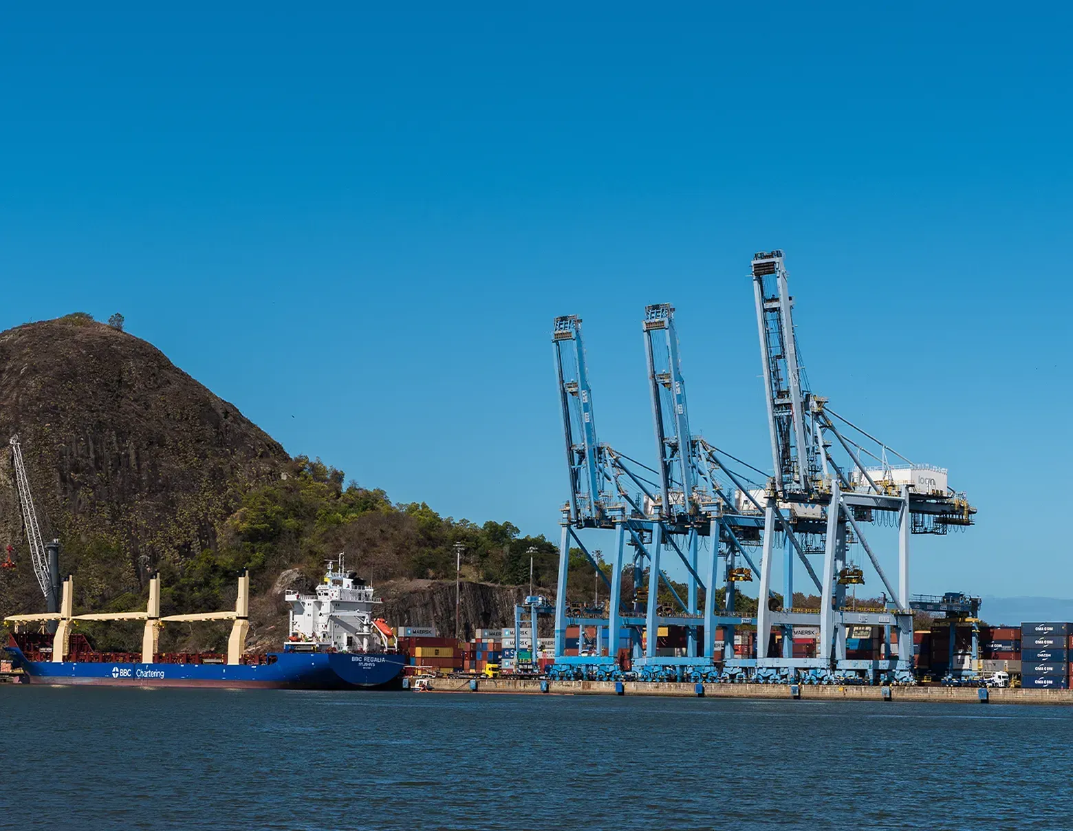 A large blue cargo ship moored at a port terminal beside three tall blue container cranes, with a grassy hill behind.