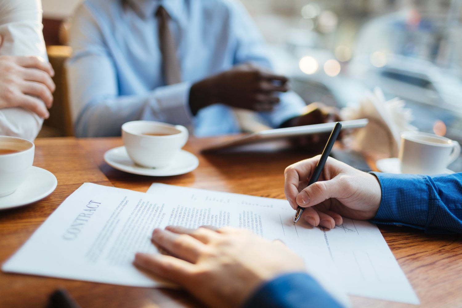 Person signing a contract at a table with two others, coffee cups, and a window in the background.