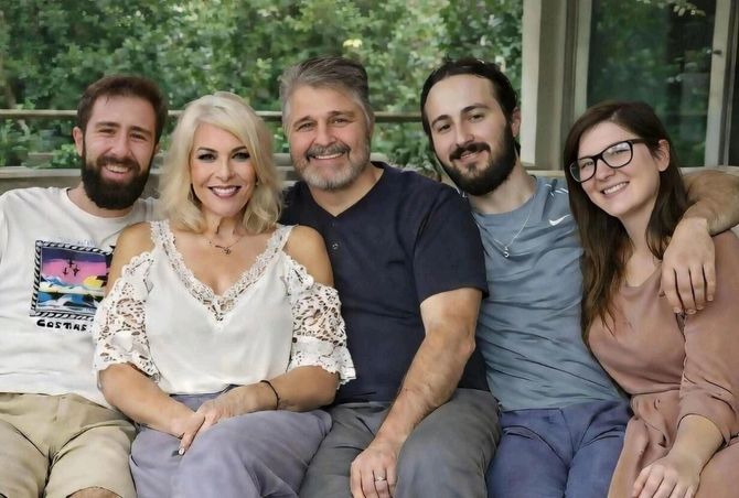Family of five smiles while sitting on a porch; greenery in the background.