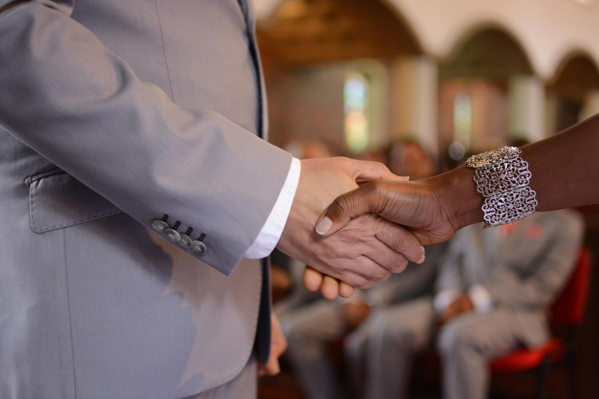 Two people shaking hands in a formal indoor setting, one in a gray suit and one in a patterned sleeve.