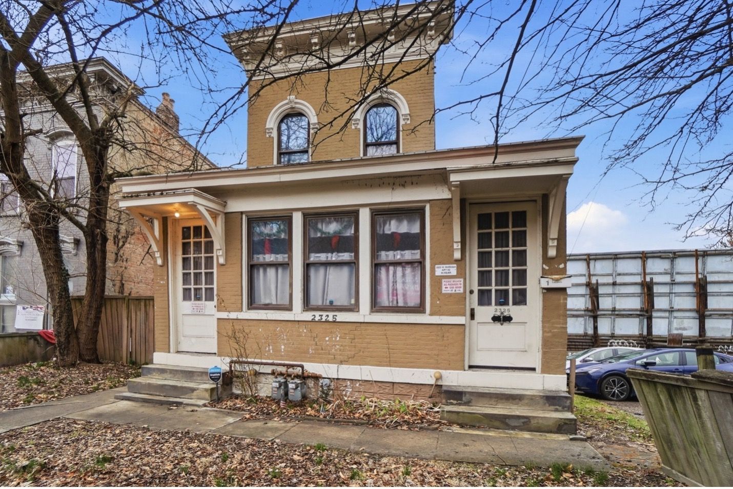 Tall white building with ornate roof and arched windows against a cloudy sky, framed by tree leaves.