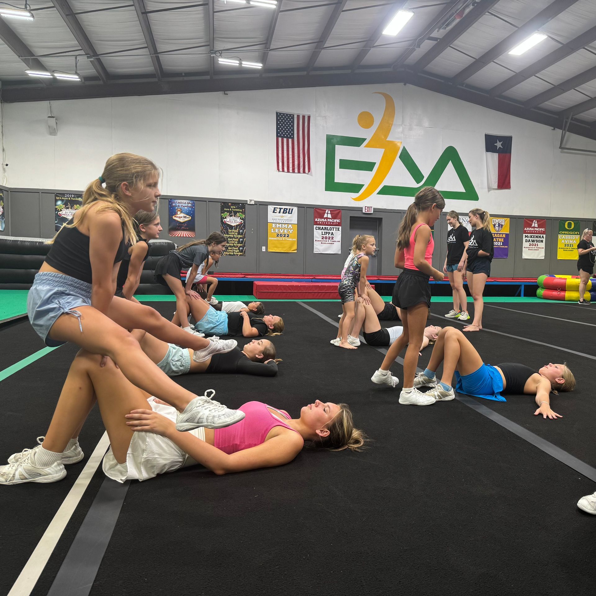 A group of girls are doing stretching exercises in a gym with the word eaa on the wall