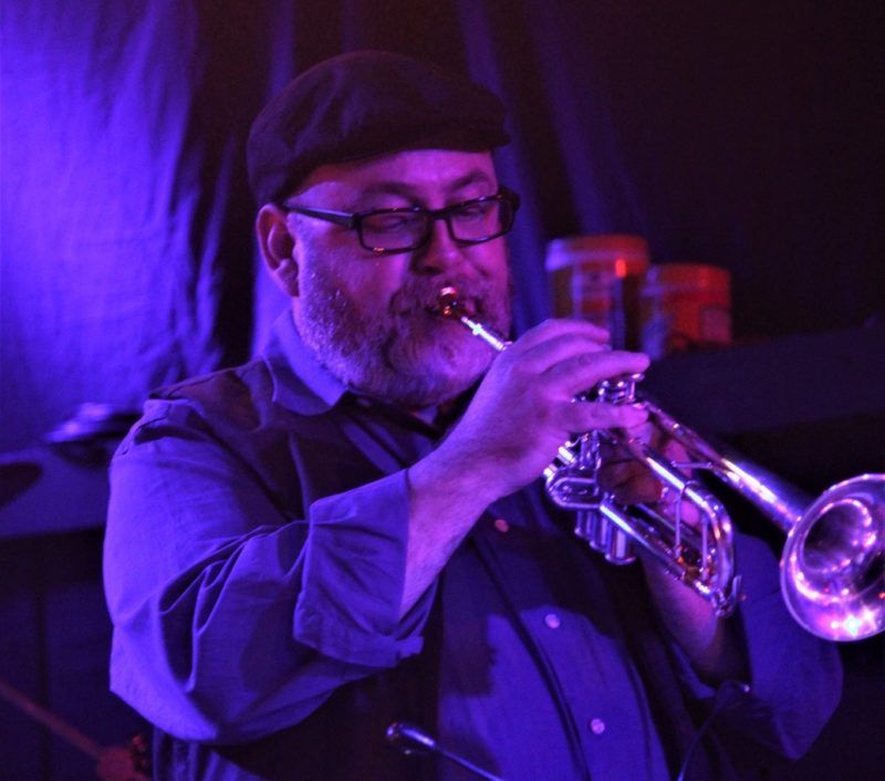 Man with glasses and a beard playing a trumpet under blue stage lights.