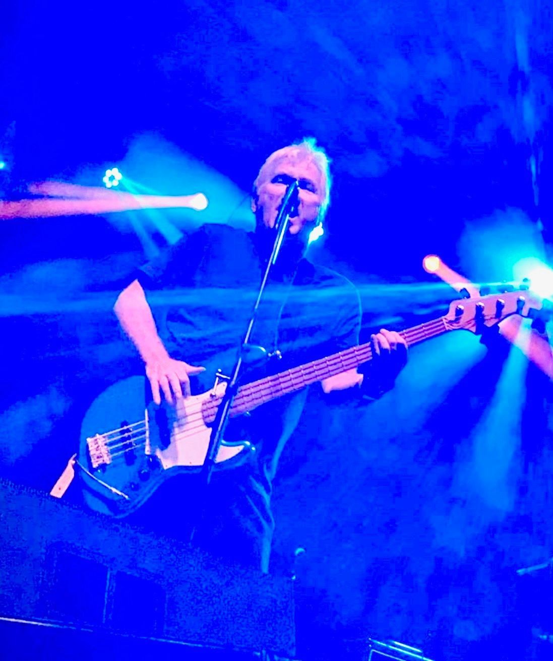Man playing a blue bass guitar on stage, lit with blue and orange lights.
