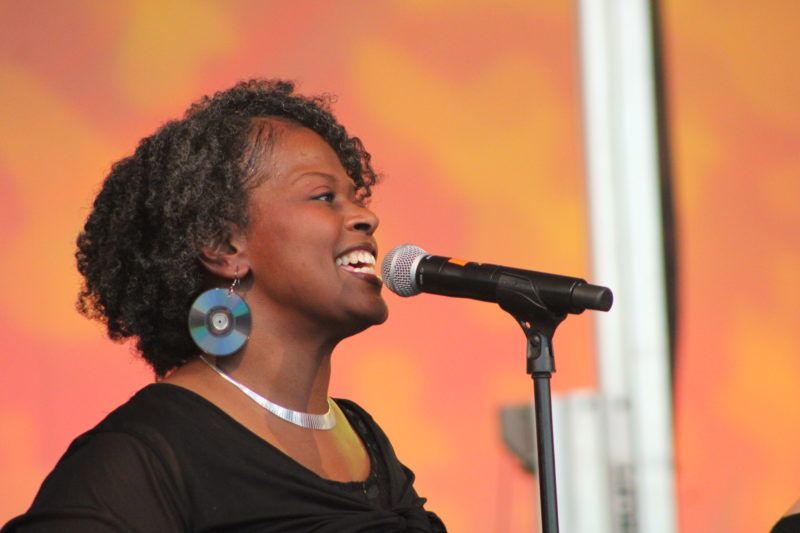 Woman with gray curly hair singing into a microphone, wearing CD earrings, on a stage with a warm orange background.