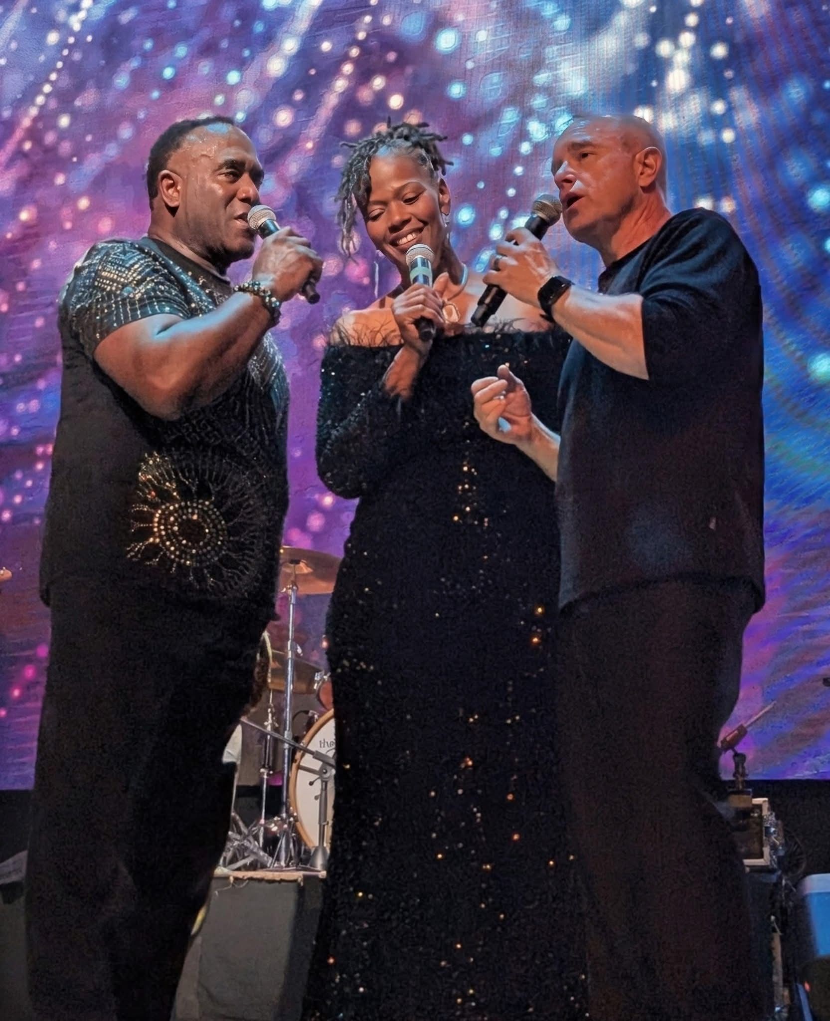 Four vocalists on stage, singing into microphones. One man wears a vest, the women are in black. Purple stage lighting.