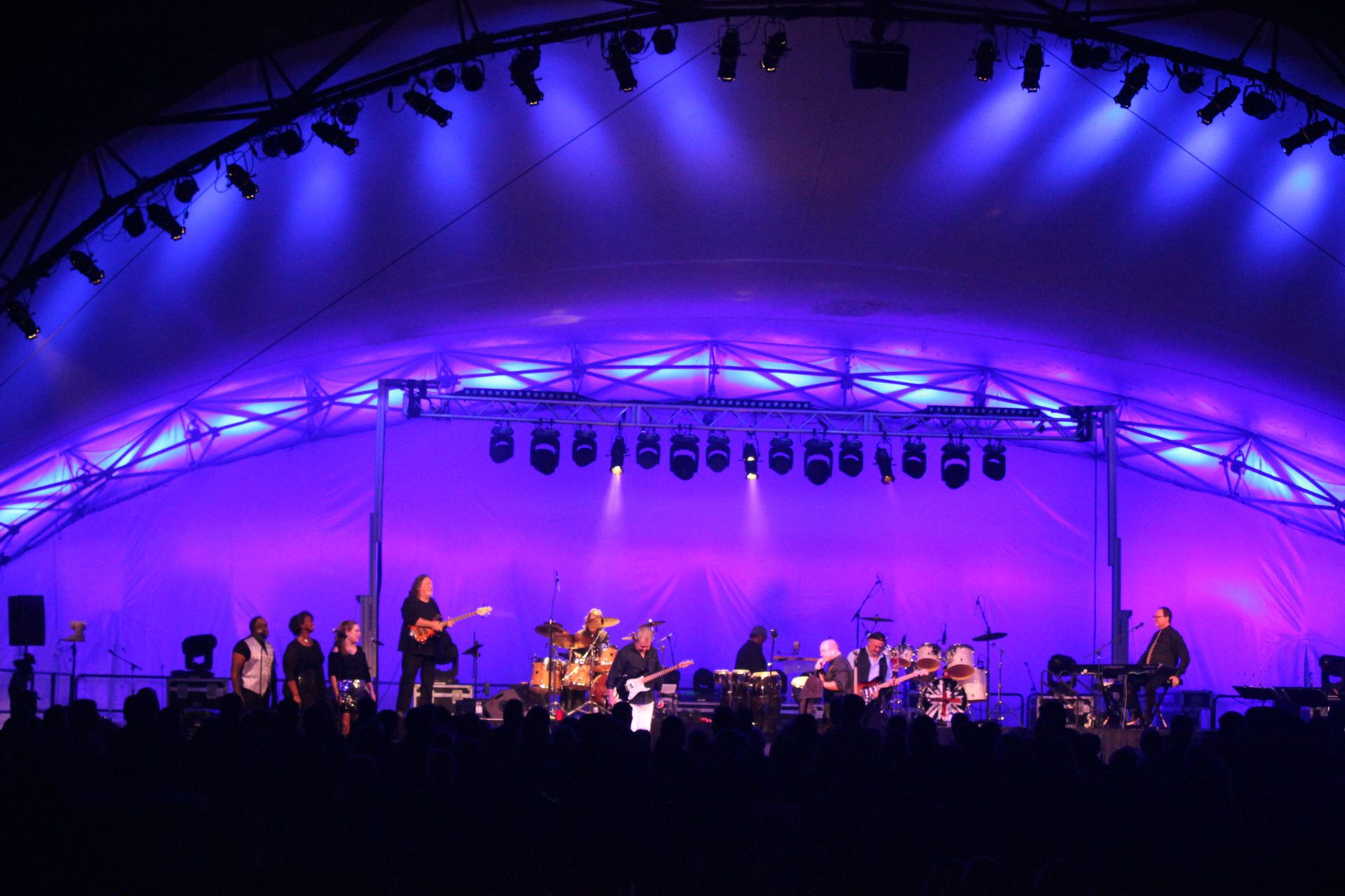 Band performing on stage under a purple-lit tent. Musicians include a drummer, guitarists, and a singer.