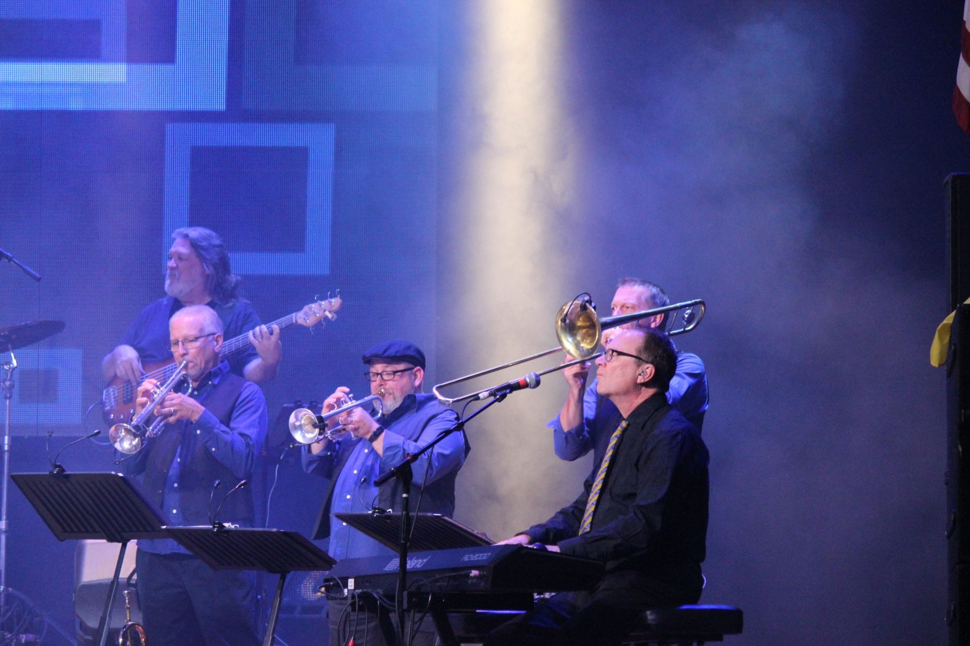Band performing on stage with brass instruments, keyboard, and guitar under blue stage lighting.