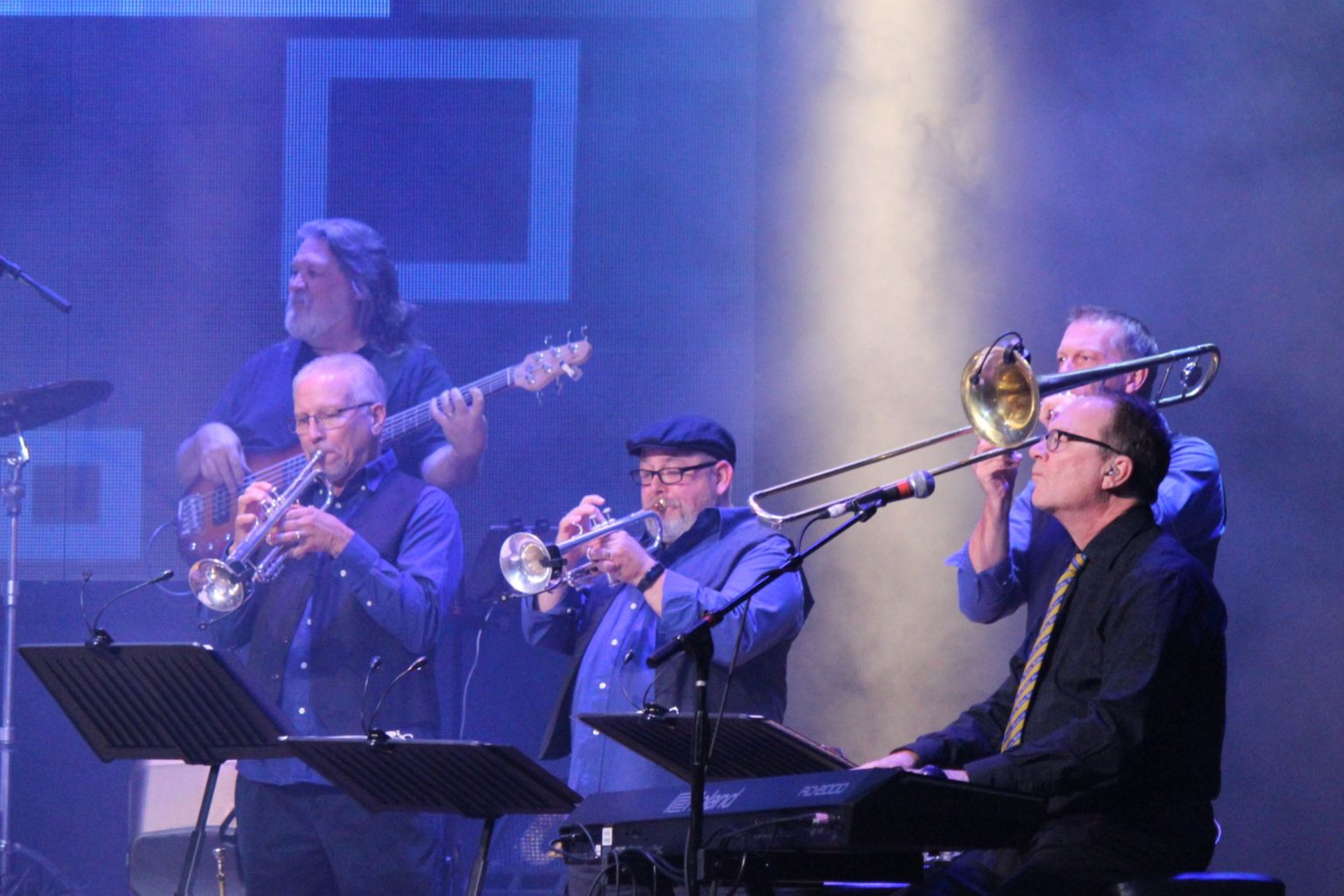 Band on stage performing, with trumpet, trombone, guitar, keys, and drums. Blue lighting.
