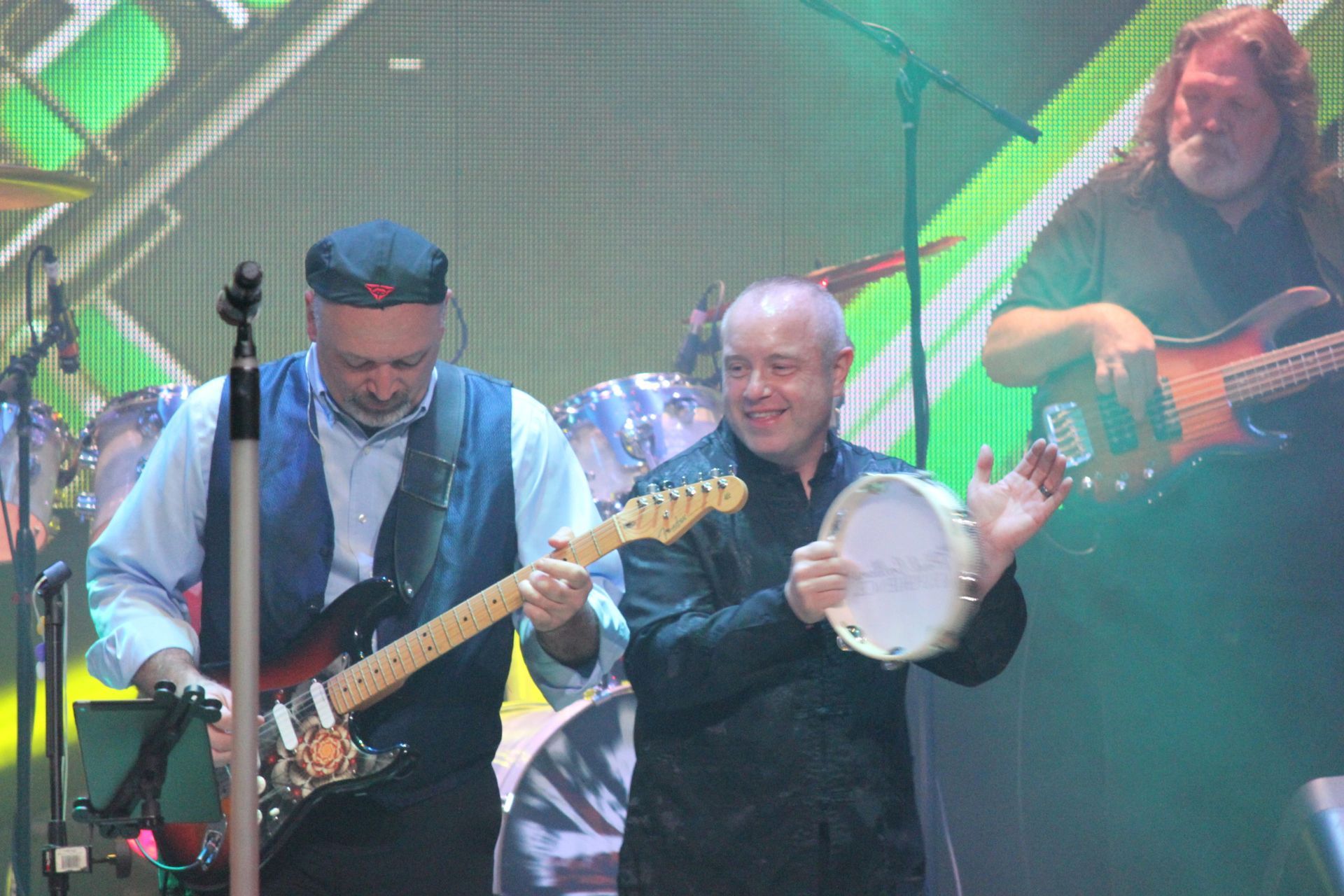 Band performing on stage. Two men in foreground; one plays guitar, the other tambourine.