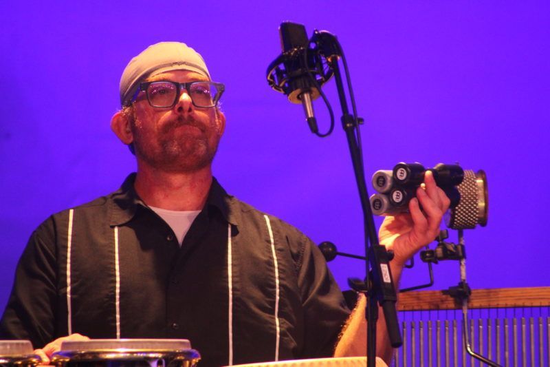 Man in glasses, beanie, and open black shirt, holding a unique audio recorder in front of a microphone; stage with blue backdrop.