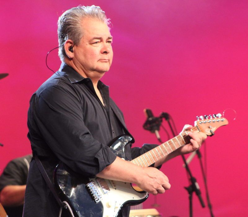 Man playing electric guitar on stage, wearing a black shirt, pink backdrop.