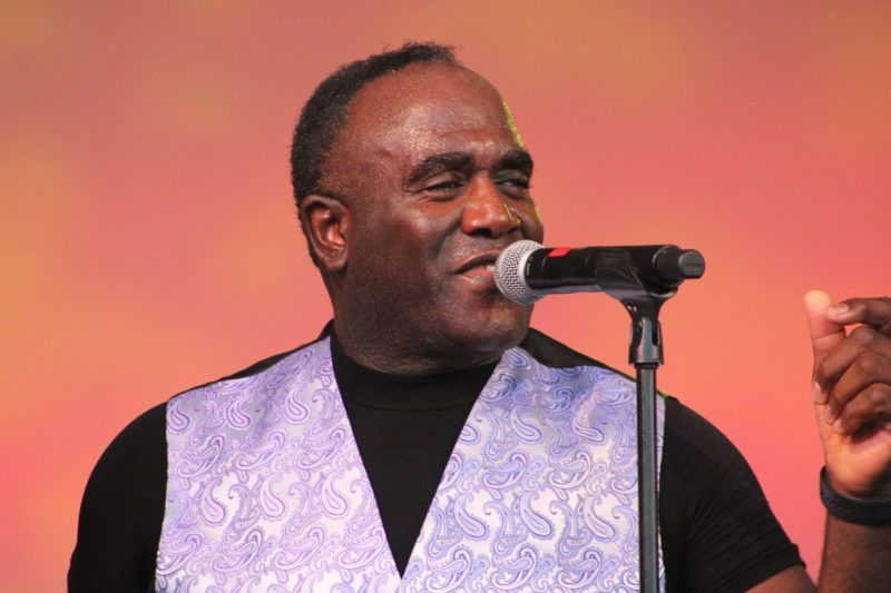 Man singing into a microphone, wearing a black shirt and paisley vest, orange background.