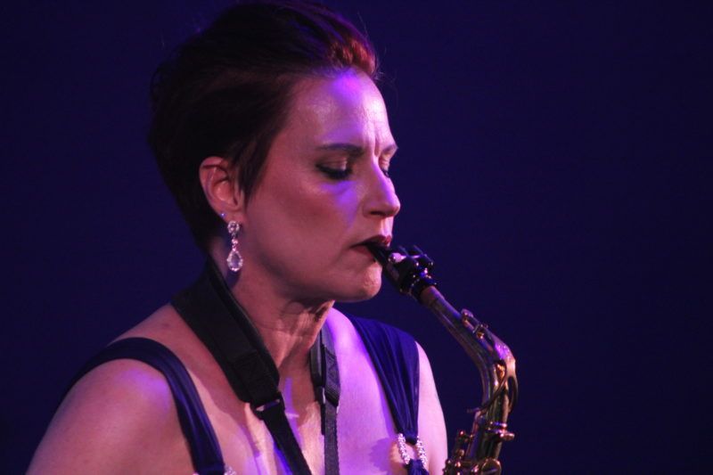 Woman playing a saxophone, dark background, blue lighting. Wearing earrings and a sleeveless dress.