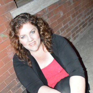 Woman with curly brown hair, smiling, wearing a red top and black cardigan in front of a brick wall.