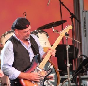 Man playing a sunburst electric guitar on stage, wearing a vest and cap, drums in the background.