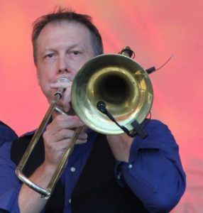 Man playing trombone, close-up; wearing blue shirt and black vest, playing at a live event.