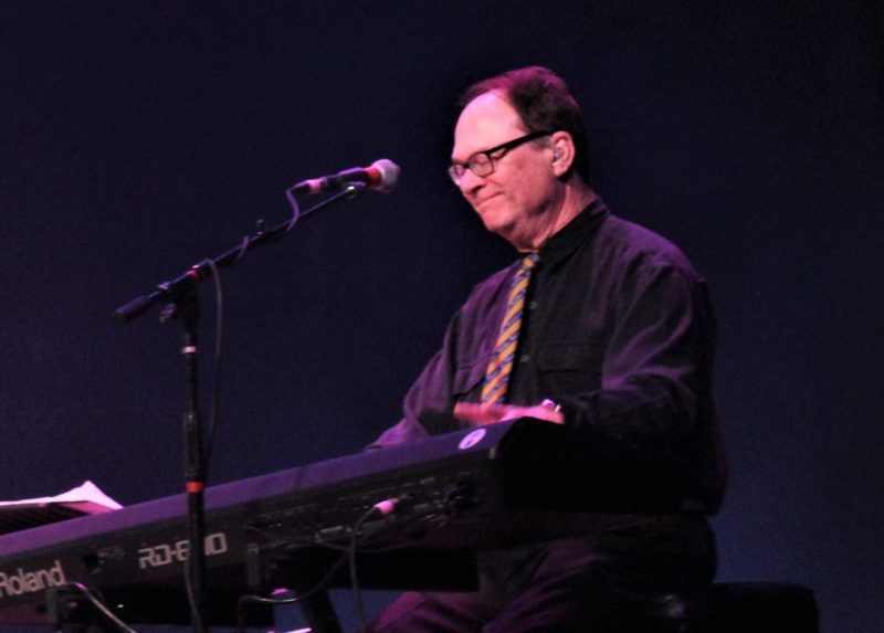 Keyboardist playing on stage, wearing glasses, black shirt, and tie. Dark background.