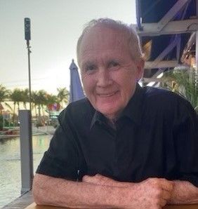 Smiling older man wearing a black shirt sits outdoors, possibly near water, with palm trees and a pier in the background.