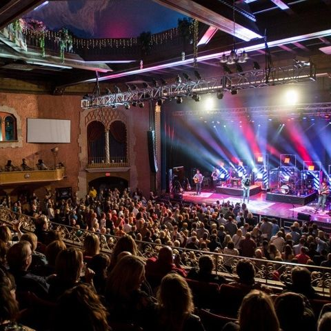 Concert in a grand theater: Band on stage, audience seated, colorful stage lighting.
