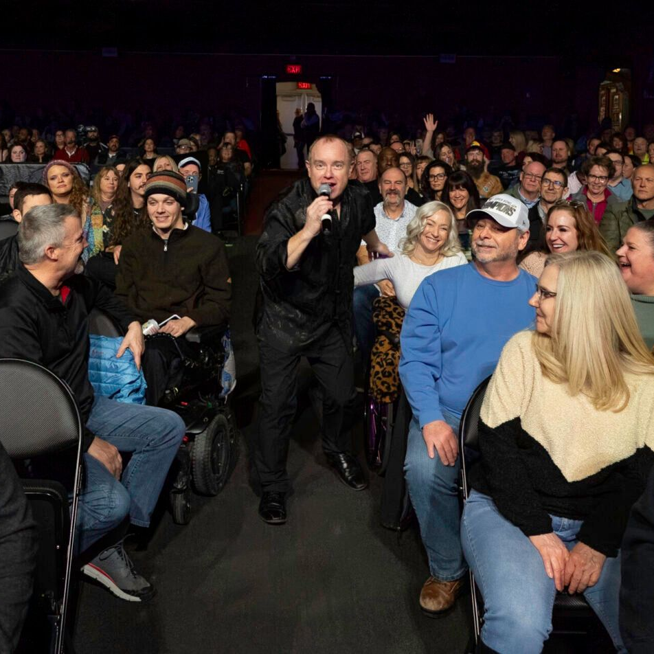 A man with a microphone performs on stage, facing a diverse, seated audience in a theater.