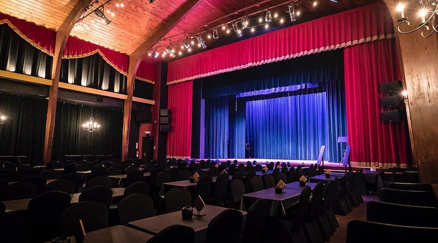 Theater with a stage and red curtains, rows of tables and black chairs.