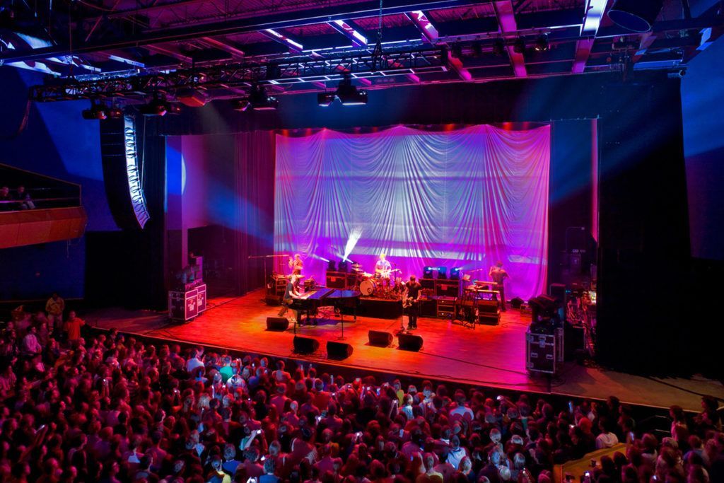 Concert stage with band and audience; purple, blue, and red lighting illuminates the scene.