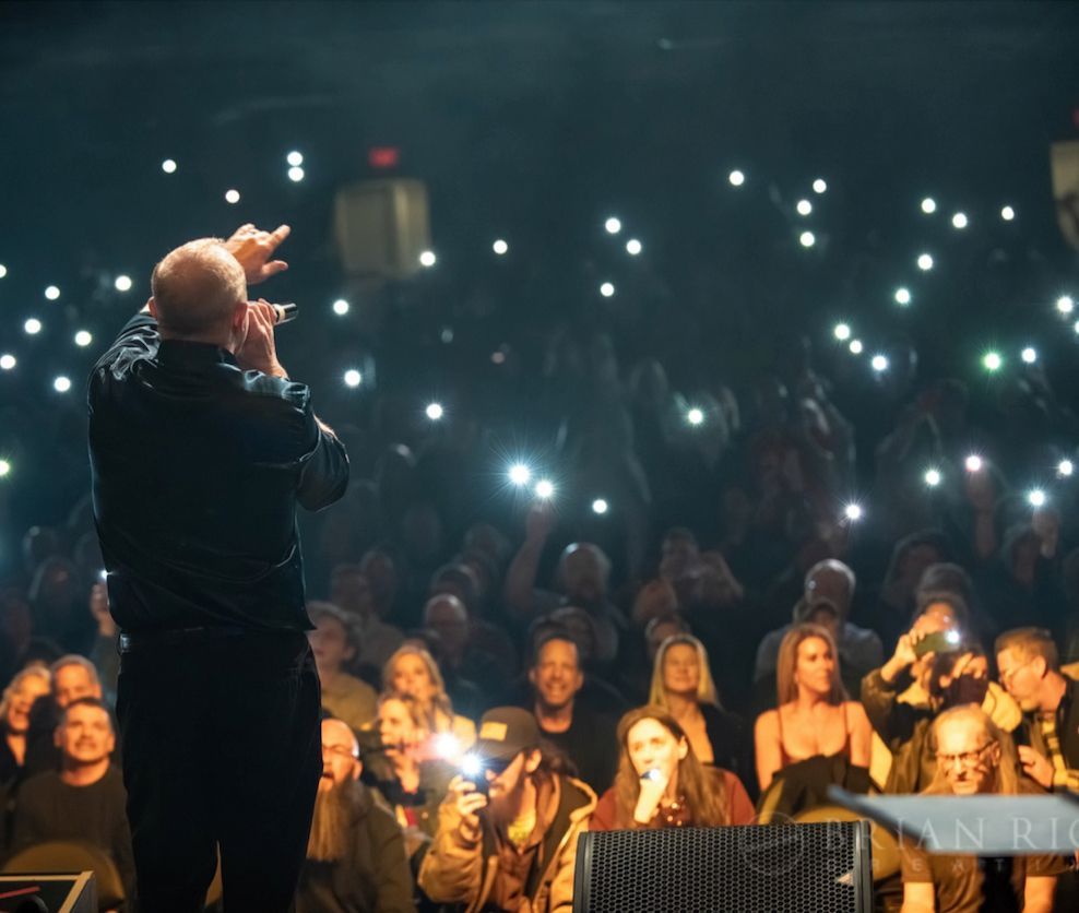 A bald man sings on stage to a crowd holding up illuminated cell phones. Dark setting.