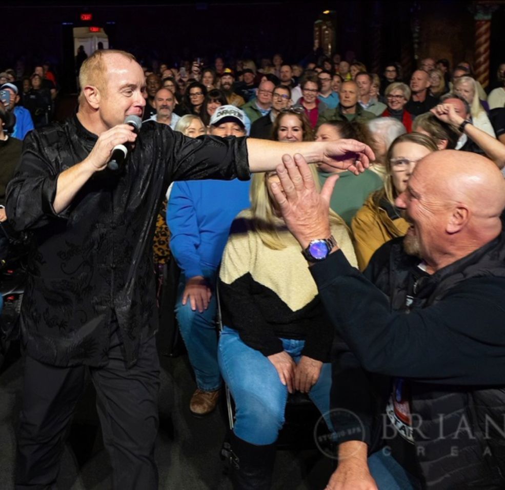 Comedian in black shirt interacts with a bald man in the audience, large crowd watches.