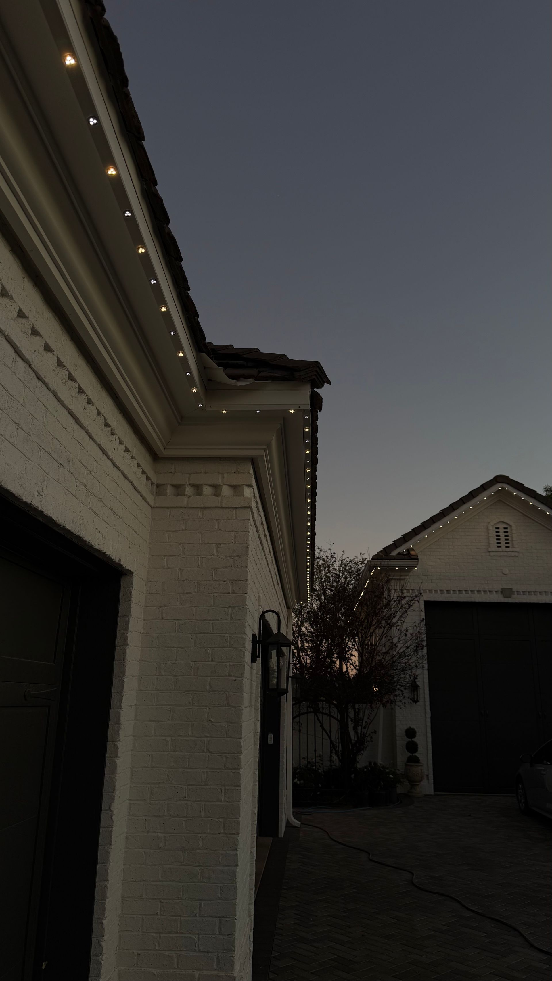 Dusk view of a white brick house with roofline lights and a driveway leading to a garage