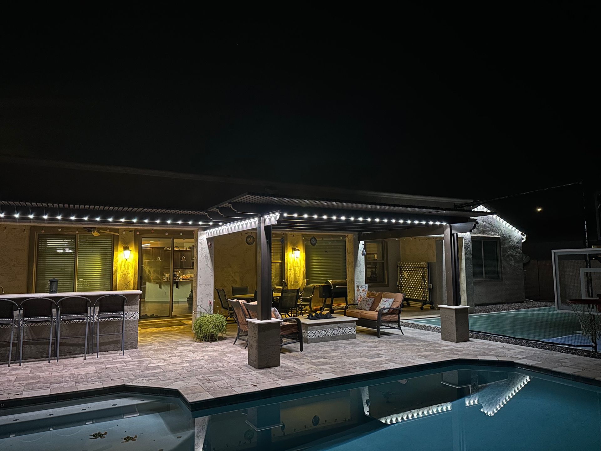 Night view of a patio with a pool. String lights illuminate the space with a bar and seating area.