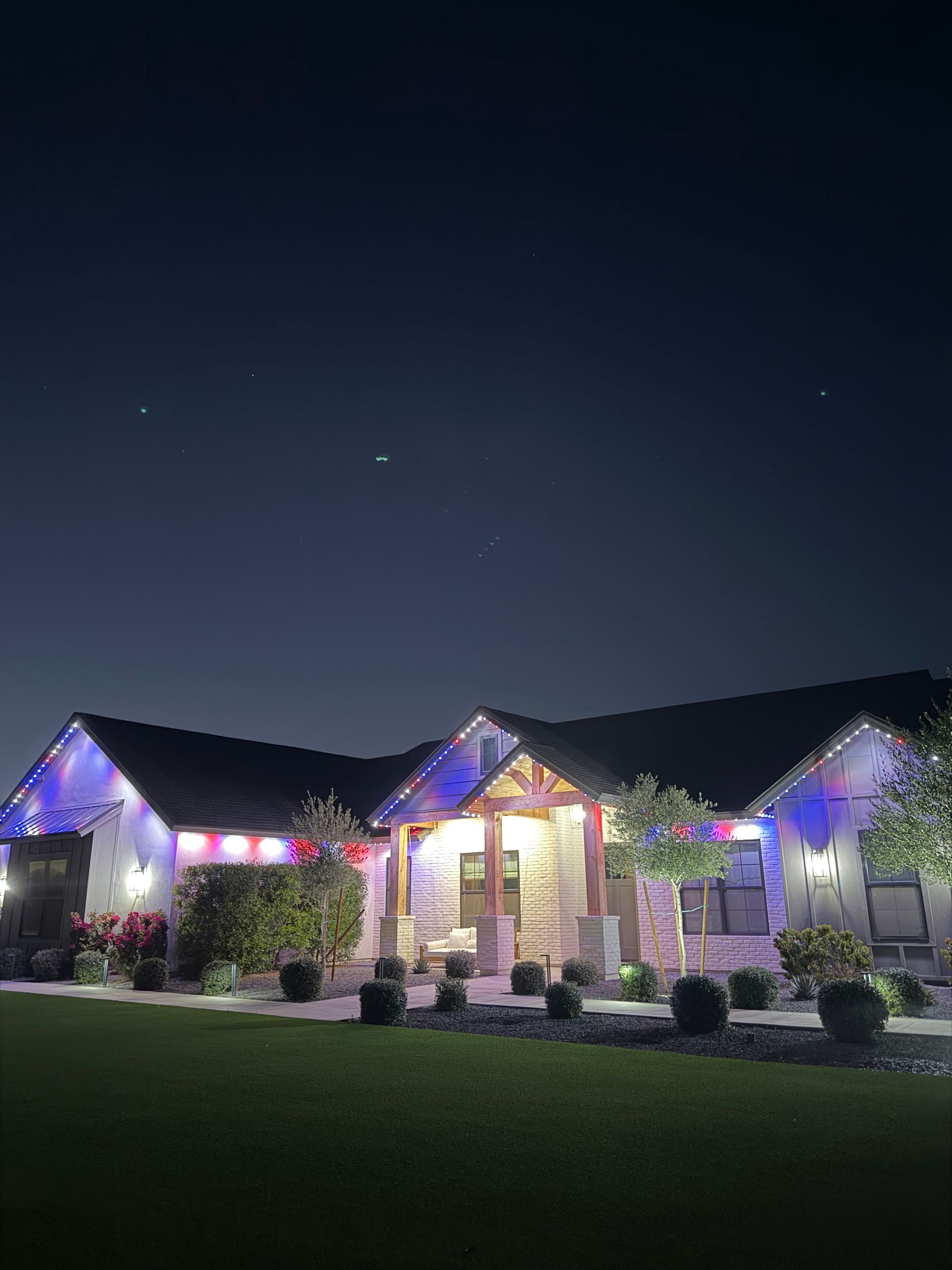 Nighttime photo of a well-lit house with purple and warm lights, front porch, and manicured lawn