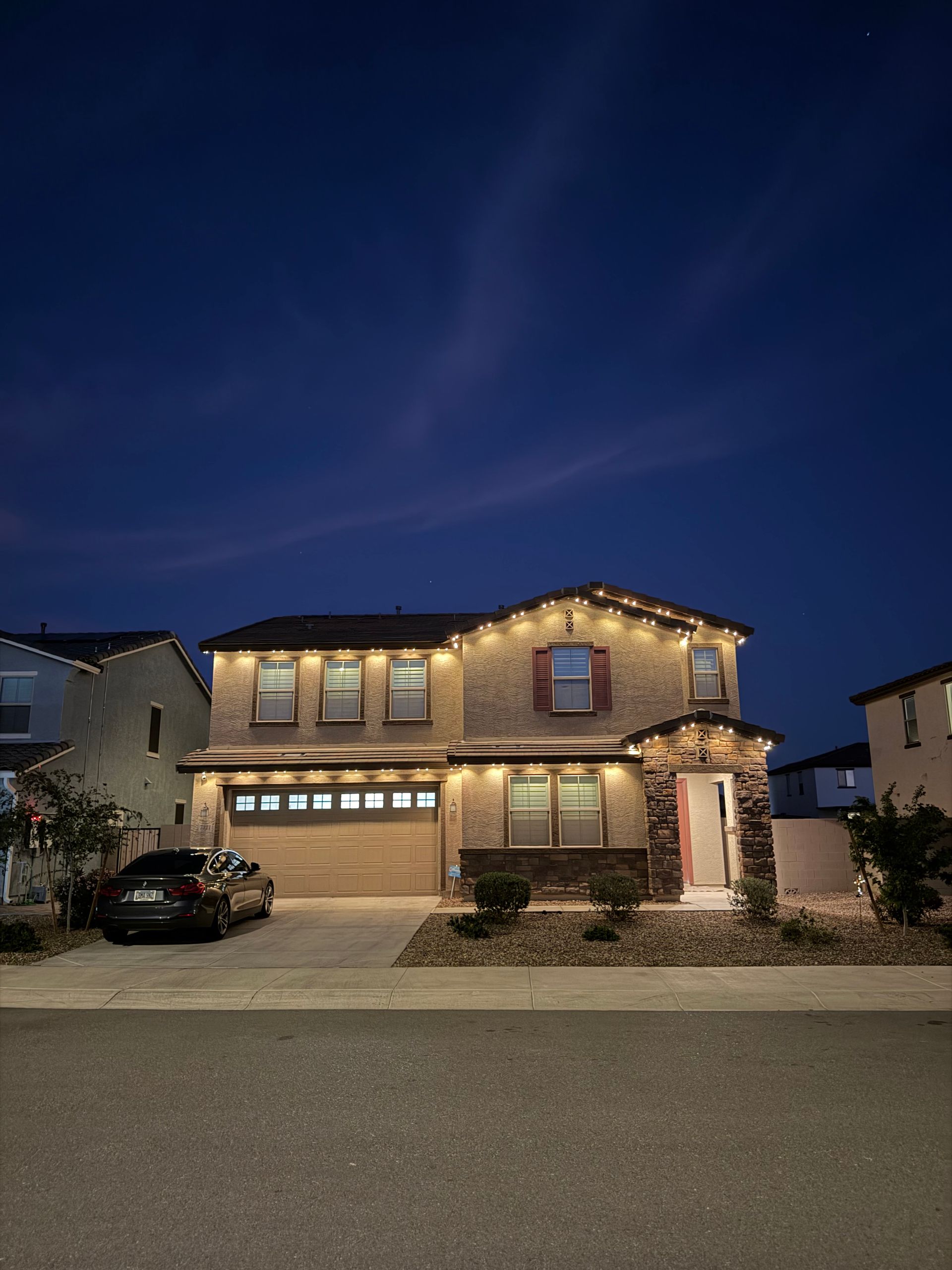 A two-story suburban house at twilight, illuminated by festive string lights along the roofline and driveway.