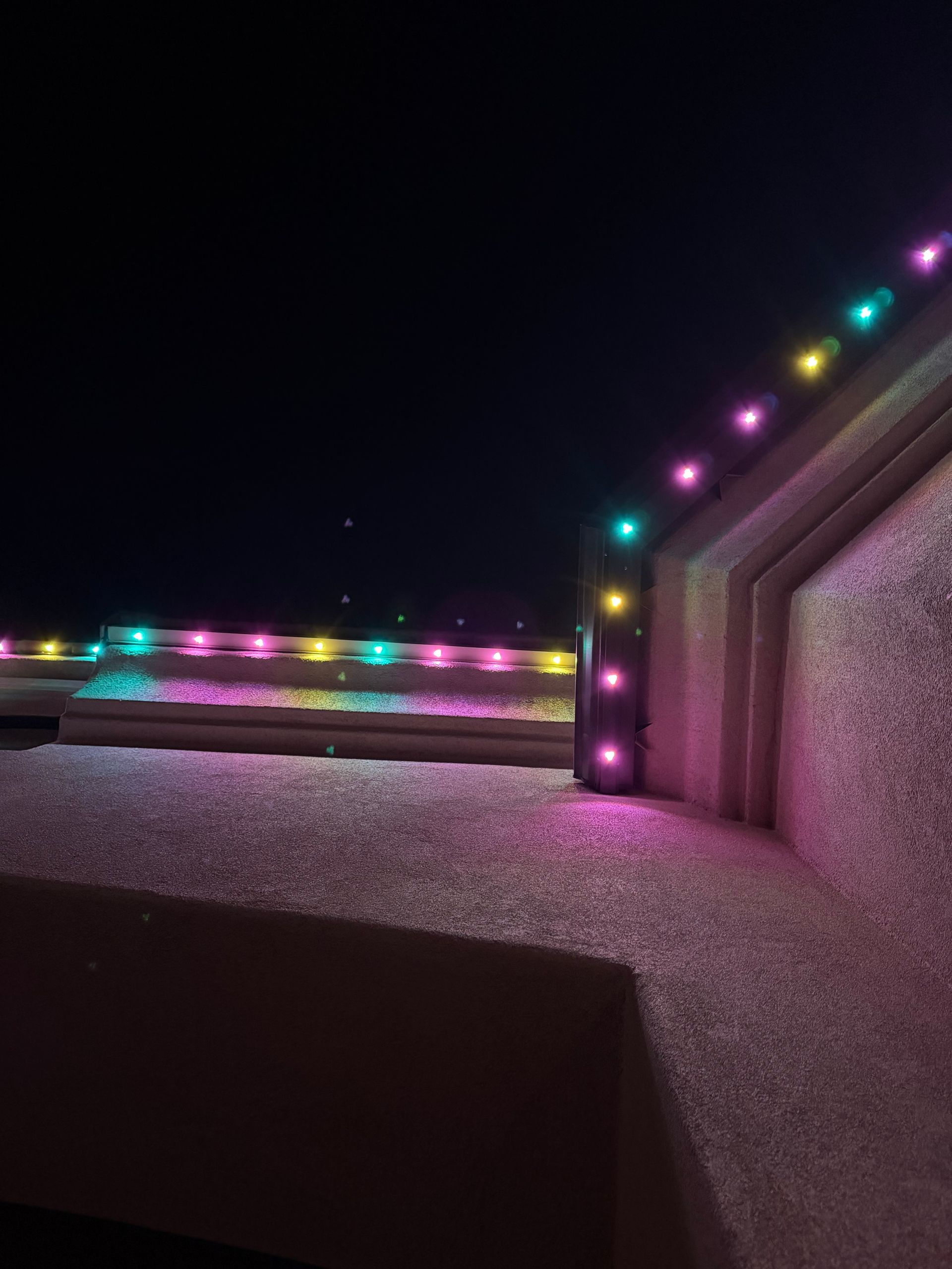Colorful LED string lights along a rooftop edge at night, glowing pink, green, blue, and yellow.
