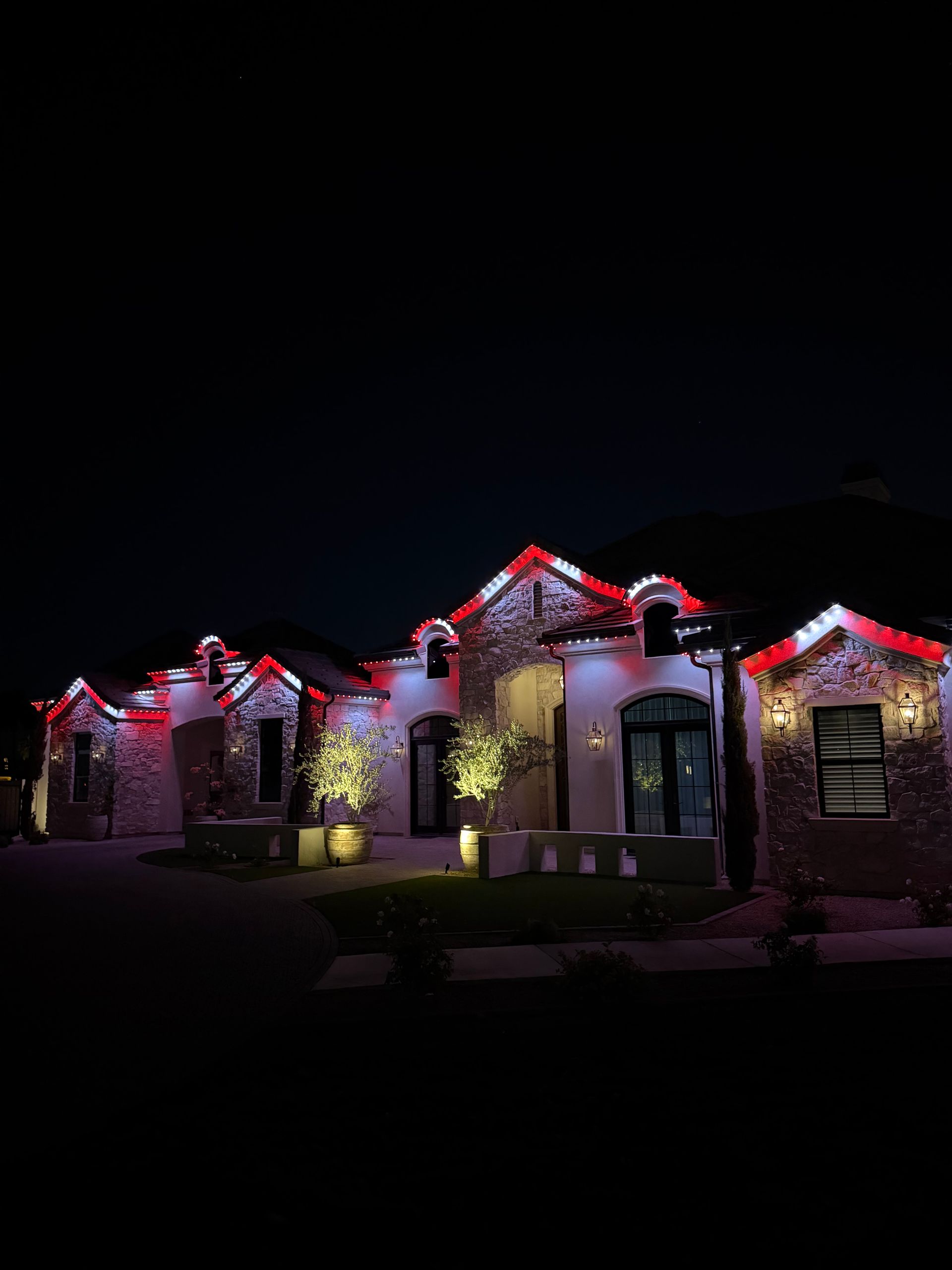 House exterior at night decorated with red roof lights and glowing white holiday lights in the yard