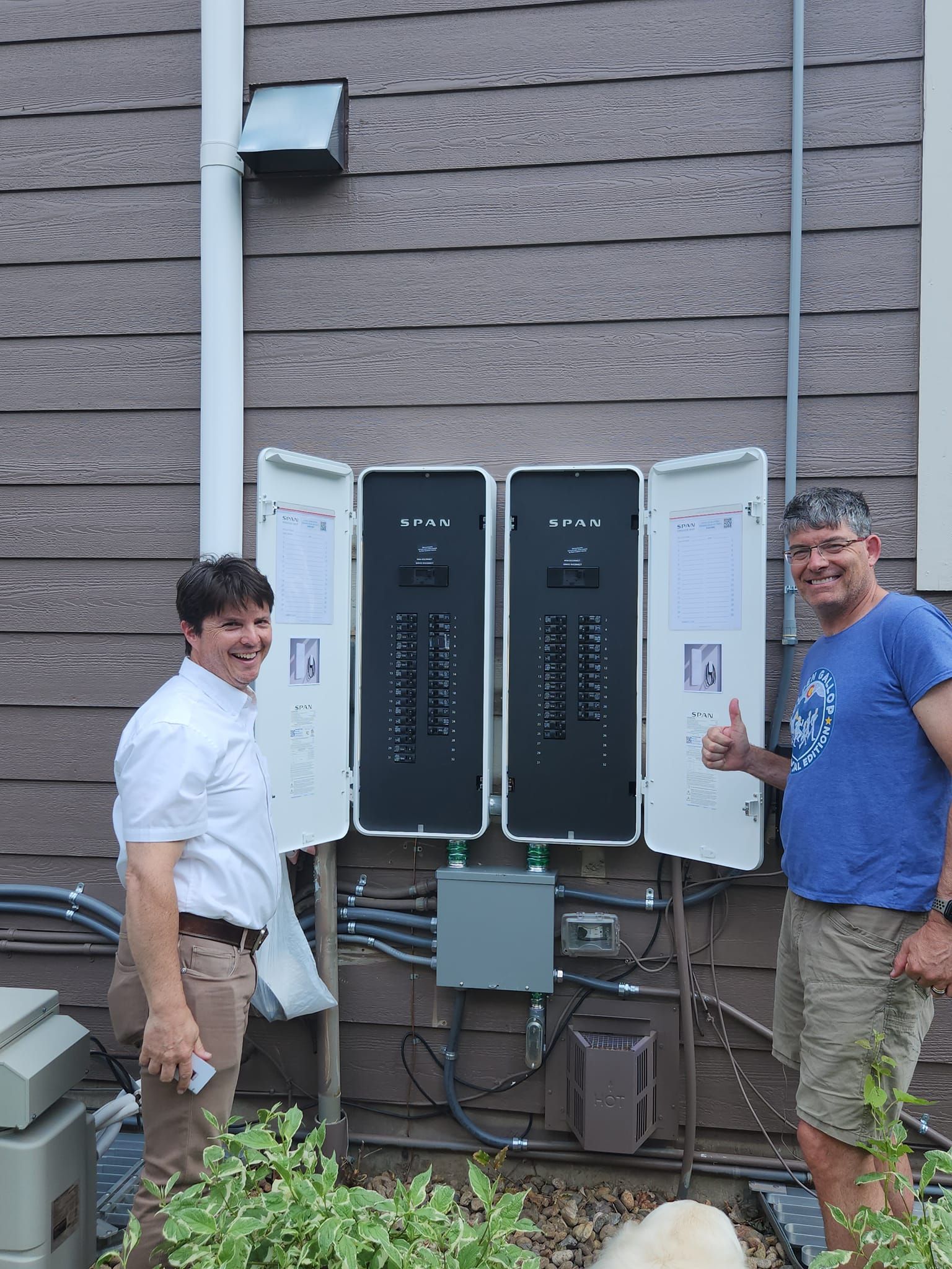 Two men are standing next to a electrical box on the side of a house.
