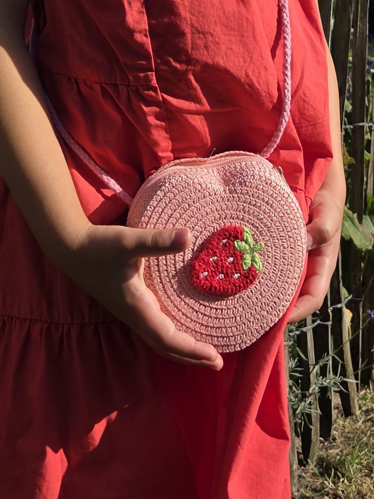 Kind met een roze tasje versierd met aardbeien, gekleed in een rode jurk, buiten.
