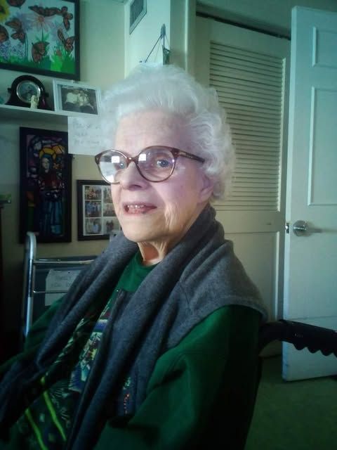 An elderly woman wearing glasses and a green shirt is sitting in a chair
